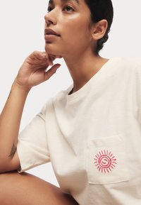 Krämfärgad bomulls-t-shirt med kort ärm, med en broderad röd sol design på vänster bröstficka. Mjuk textur och avslappnad passform.