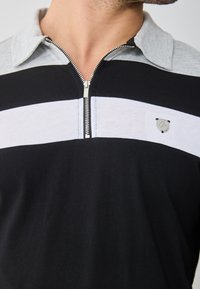 Černé a šedé polo tričko s polovičním zipem, které má vodorovné pruhy, stříbrný zip a malou nášivku s logem na hrudi. Hladký bavlněný materiál.