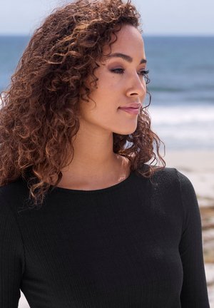 Schwarzes, geripptes Langarmoberteil mit einem runden Ausschnitt. Das Model hat lockige Haare und einen Strandhintergrund mit sanft unscharfem Ozean.