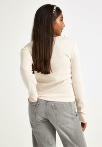 Ung kvinde med langt mørkt hår iført en beige langærmet bluse, grå jeans og et leopardmønstret pandebånd, stående med ryggen til.