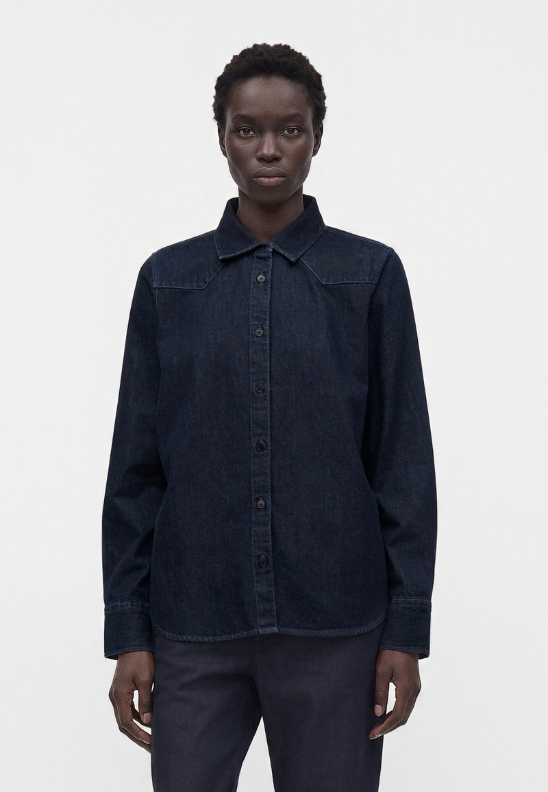 Personne portant une chemise en denim sombre à manches longues et col, se tenant devant un fond blanc uni.