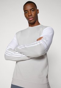 Sweatshirt gris clair avec des manches blanches, col ras du cou et trois rayures blanches le long des bras. Texture lisse avec une coupe ajustée.
