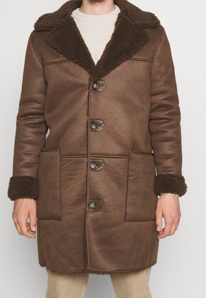 Man draagt een mid-lange bruine schapenvachtjas met grote kraag, knoopsluitingen, opgestikte zakken en omgeslagen mouwen over een lichte trui.