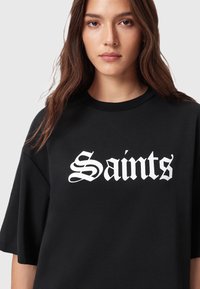 Zwarte T-shirt met korte mouwen, voorzien van vet witte gotische letters die "Saints" over de voorkant zeggen. Zachte, gladde textuur.