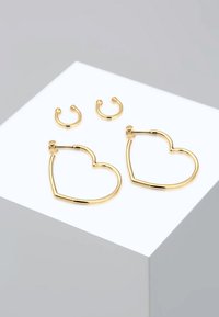 Pendientes de aro en forma de corazón dorado y dos pequeños pendientes de puño dorados sobre un soporte gris claro. Superficie lisa, acabado brillante, diseño minimalista.
