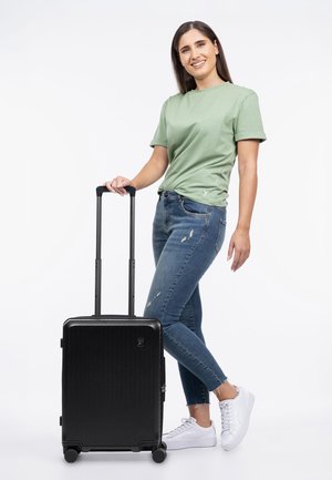 Donna con t-shirt verde e jeans in piedi con una valigia a rotelle nera, sorridendo, su uno sfondo bianco uniforme.