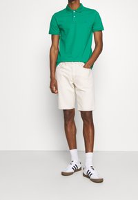Polo shirt vert avec col, shorts crème et baskets blanches avec rayures navy. Tissu lisse et coupe décontractée.