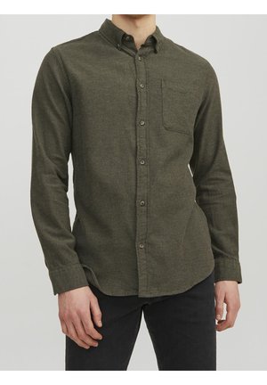 Homme portant une chemise kaki boutonnée avec une poche poitrine et des manches longues, associée à un pantalon noir.
