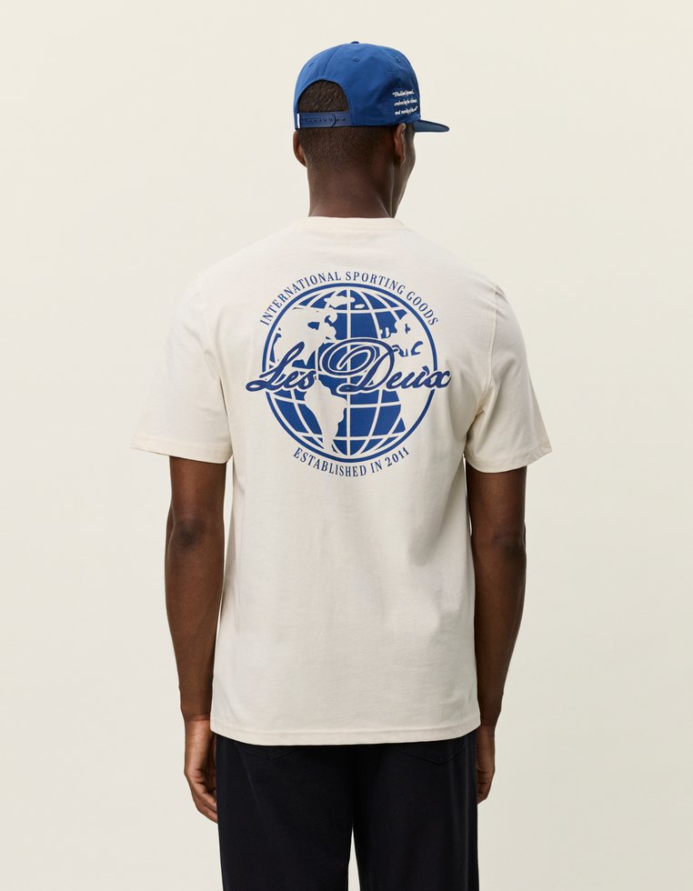 Beżowa bawełniana koszulka z niebieską grafiką globusa na plecach, tekst "Les Deux" oraz szczegóły "International Sporting Goods Established in 2011".