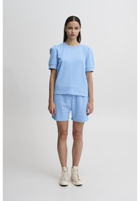 Top corto in cotone azzurro chiaro con maniche a sbuffo, abbinato a pantaloni corti coordinati. Il modello indossa sneaker alte beige. Lo sfondo è semplice.