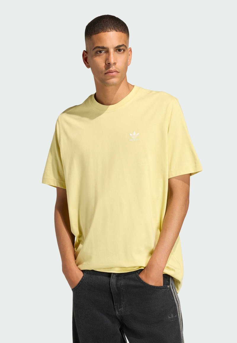 Yellow cotton t-shirt with short sleeves, round neckline, and white logo detail on the chest. Paired with dark denim jeans.
