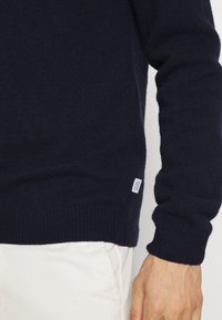Pulover tricotat de culoare albastru marin, cu manșete și tiv cu rigle, având o mică etichetă a mărcii pe partea inferioară. Textură moale, design casual.