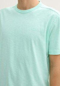 Hombre vistiendo una camiseta de cuello redondo de manga corta en un azul turquesa claro con un pequeño logo en relieve y "1962" en el pecho.