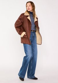Veste en cuir marron avec un col en shearling beige, associée à un jean large bleu clair. Des chaussures noires complètent le look.
