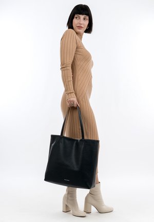 Black leather tote bag with a smooth texture, featuring two sturdy handles and a spacious interior. The model wears a ribbed beige dress.