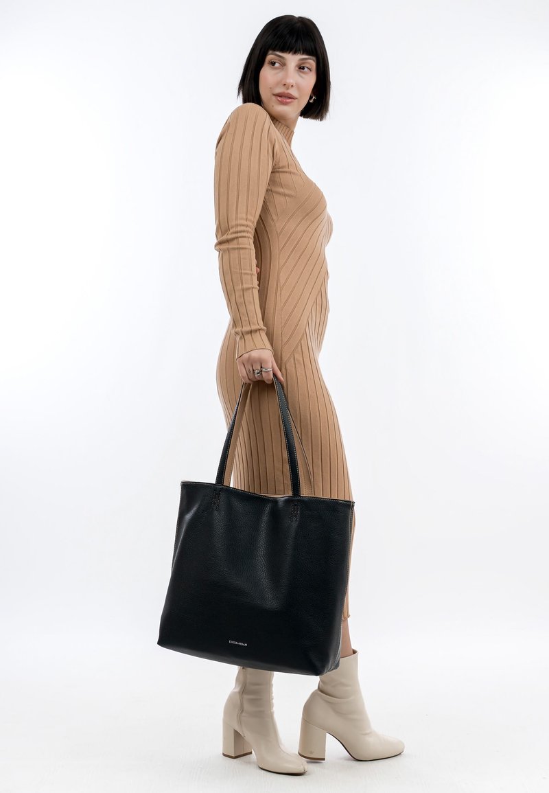 Sac fourre-tout en cuir noir avec une texture lisse, doté de deux poignées robustes et d'un intérieur spacieux. Le modèle porte une robe beige à côtes.