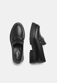 Mocassins en cuir noir avec une semelle épaisse, dotés d'une sangle décorative et de détails en métal. Texture lisse et conception à bout arrondi.