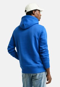 Blauwe hoodie met een trekkoord in de capuchon, ribgebreide manchetten en onderrand. De stof lijkt glad en dik, gezien van achteren. Gekocht met lichtblauwe jeans.