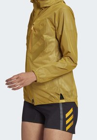 Lichtgewicht gele jas met capuchon, ritssluiting aan de voorkant en een meshstructuur. Gecombineerd met zwarte shorts met gele en grijze accenten.