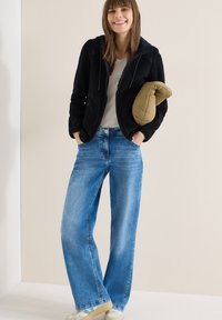 Femme souriante portant une chemise beige, une veste noire zippée, un jean bleu à jambes larges, tenant une veste olive pliée, debout devant un fond clair uni.