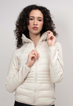 Mujer con cabello rizado cerrándose una chaqueta acolchada beige claro con cuello alto, sobre un fondo liso.