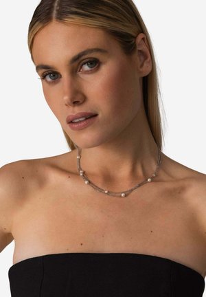 Frau mit glattem blondem Haar, die ein schwarzes trägerloses Top und eine mehrlagige silberne Halskette mit kleinen Perlen trägt.