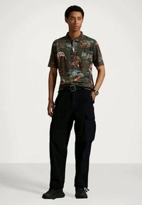 Joven con una camiseta polo verde oscuro con estampados de animales, pantalones cargo negros, zapatillas negras y un reloj de pulsera, de pie con una mano en el bolsillo.