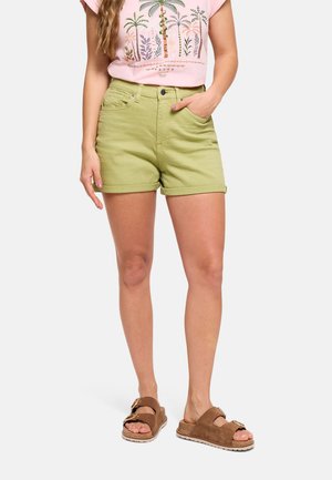 Mujer que lleva pantalones cortos verde claro con dobladillo enrollado, camiseta rosa con estampado de palmera y sandalias marrones de doble tira, de pie con una mano en el bolsillo.