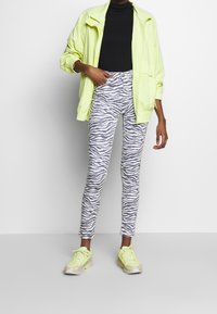 Persona vestida con una chaqueta de neón amarilla, camiseta negra, pantalones de estampado de cebra en gris y blanco, y zapatillas de neón amarillas, de pie contra un fondo blanco.