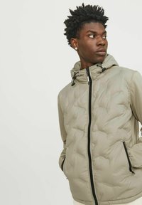 Beige pufferjack met een rits aan de voorkant, aantrekbare capuchon en zijzakken. Heeft een gequiltde textuur met zwarte ritsaccenten.