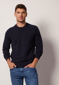 Marinblå stickad tröja med rund hals, ribbade ärmslut och nederkant. Tyget verkar mjukt och texturerat, matchat med ljusblå jeans.