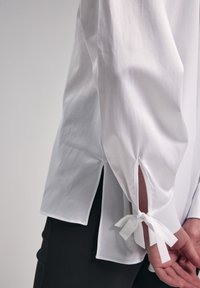 Camicia bianca a maniche lunghe con fessure laterali e nastri di stoffa ai polsini, realizzata in cotone morbido. Design semplice e aderente.