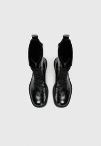 Bottes de combat en cuir noir avec une finition texturée, dotées d'un long lacet tressé et d'un bout rond lisse. Fermeture éclair sur le côté.