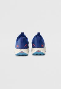 Une paire de chaussures de sport bleues avec des semelles intermédiaires blanches tachetées et des logos triangulaires rouges, vues de l'arrière sur un fond uni.