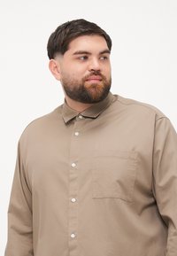 Beige Hemd mit spitzen Kragen, einer einzelnen Brusttasche und sichtbaren Knöpfen. Glatte Stoffqualität mit lässiger Passform.