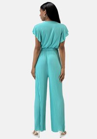 Tyrkis plisserad jumpsuit med vågiga ärmar, midja som är insvängd, vid bendesign och mjukt, lätt tyg. Visas från baksidan.