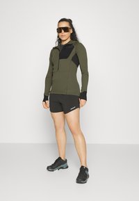 Olive green long-sleeve top with a black chest pocket, paired with black shorts and black-and-teal sneakers. Large brown sunglasses.