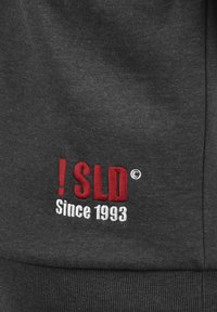 Tamno sivi dukserica s crvenim i bijelim izvezenim tekstom: "! SLD" i "Od 1993." Tkanina je meka s glatkom teksturom i rebrastim manžetama.