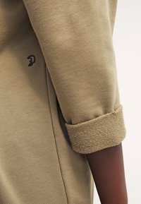 Sudadera tipo pulóver de color caqui con puños acanalados, tejido texturizado y un pequeño logo negro en la manga. Presenta un diseño de ajuste holgado.