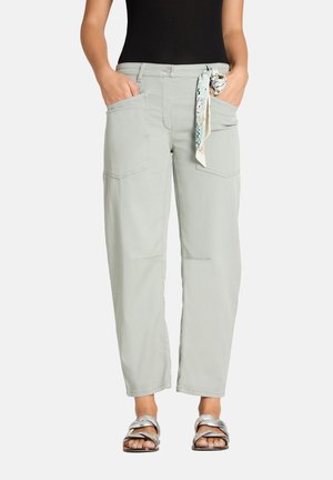 Woman wearing light gray wide-leg pants with front pockets, a patterned scarf tied at the waist, black top, and silver sandals. Hands in pockets.