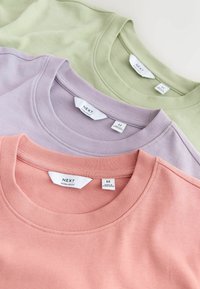 Trois t-shirts en coton pliés, dans des tons pastel rose, lavande et vert clair, chacun avec un col rond et des étiquettes de taille et de marque visibles.