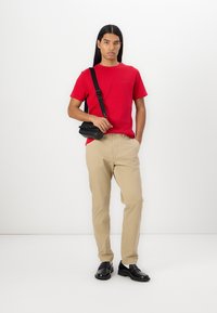 Camiseta roja de manga corta, pantalones beiges, bolsa cruzada negra y zapatos negros. Tejido suave, ajuste casual, exhibido sobre un fondo blanco.