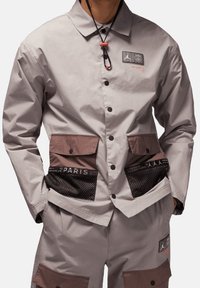 Lichtgrijze jas met bruine accenten, mesh zakken en rubberen logo patch. Voorzien van een kraag, knopen en een verstelbare koord.