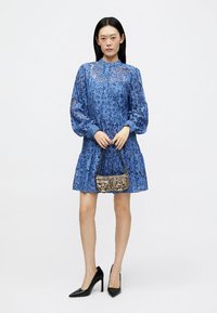 Ανοιχτό μπλε φόρεμα δαντέλας με μακριά φουσκωτά μανίκια και φουσκωτό φούστα. Το μοντέλο κρατά μια μικρή τσάντα με σχέδιο. Μαύρες γόβες ψηλοτάκουνες ολοκληρώνουν την εμφάνιση.