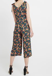 Combinaison florale avec un fond marine, présentant des motifs vifs orange, rose et vert. Liens aux épaules, jambes larges et cropped. Talons noirs.