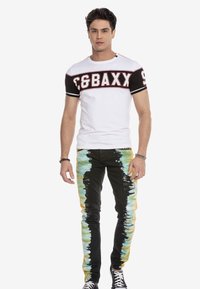 Witte t-shirt met zwarte mouwen en rode print. Zwarte jeans met een tie-dye patroon in blauw en geel. Zwarte sneakers met witte veters.