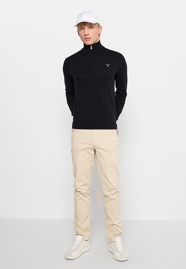 Cardigan zip nera, chinos beige, sneakers bianche e cappellino bianco. Design semplice, materiale in cotone, forma aderente, dettagli di branding minimi.