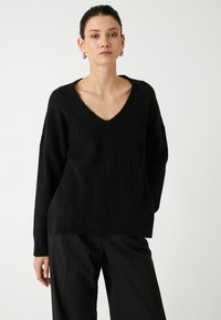 Mustassa neulotussa neuleessa on V-aukkoinen kaula-aukko, ylisuuri malli ja ribbireunus. Yhdistetty löysiin mustiin housuihin.