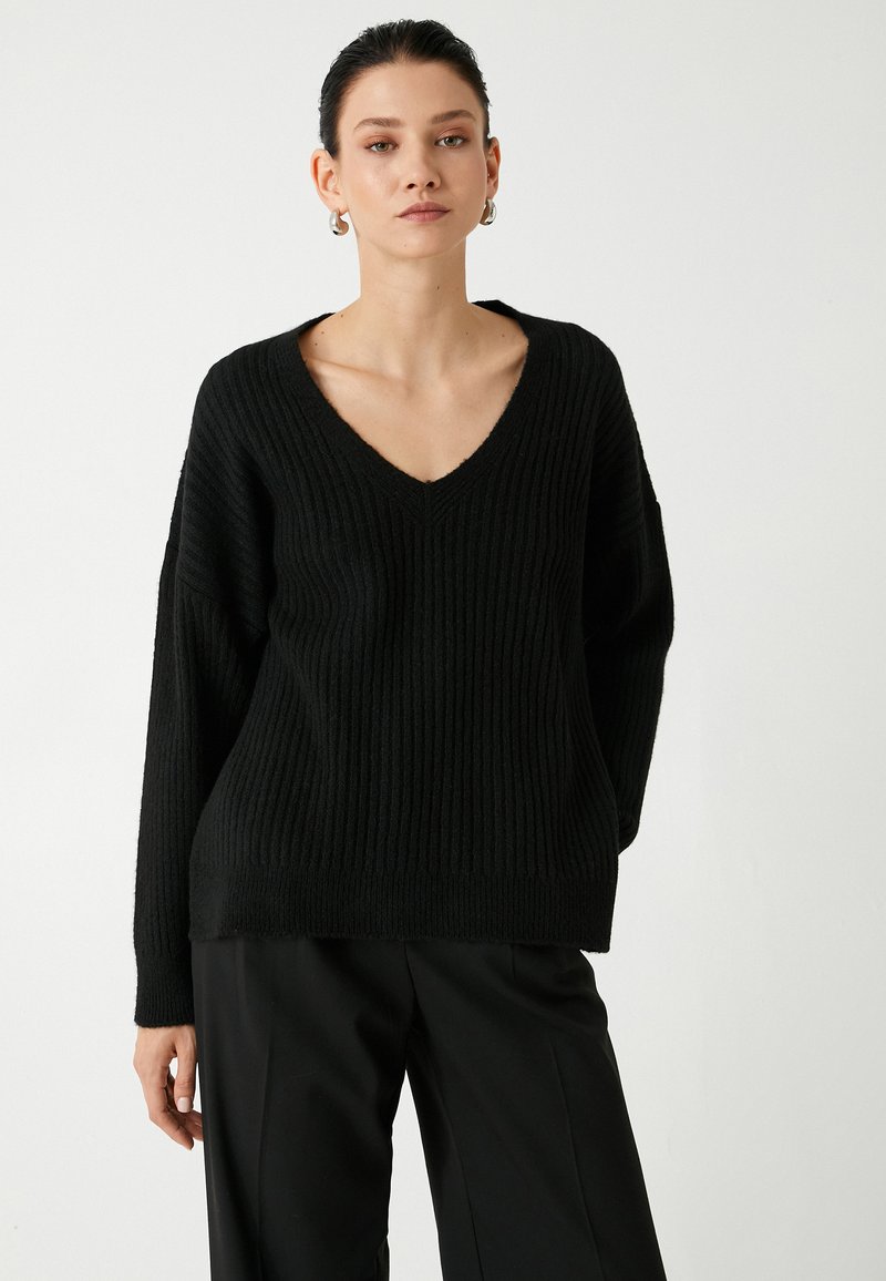 Mustassa neulotussa neuleessa on V-aukkoinen kaula-aukko, ylisuuri malli ja ribbireunus. Yhdistetty löysiin mustiin housuihin.