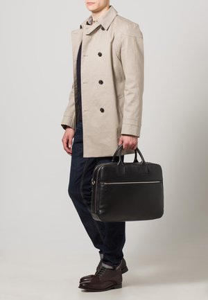 Trench coat beige doppiopetto sopra una maglia scura, jeans blu scuro, scarpe in pelle marrone, mentre tiene una valigetta in pelle nera con tasca zippata.